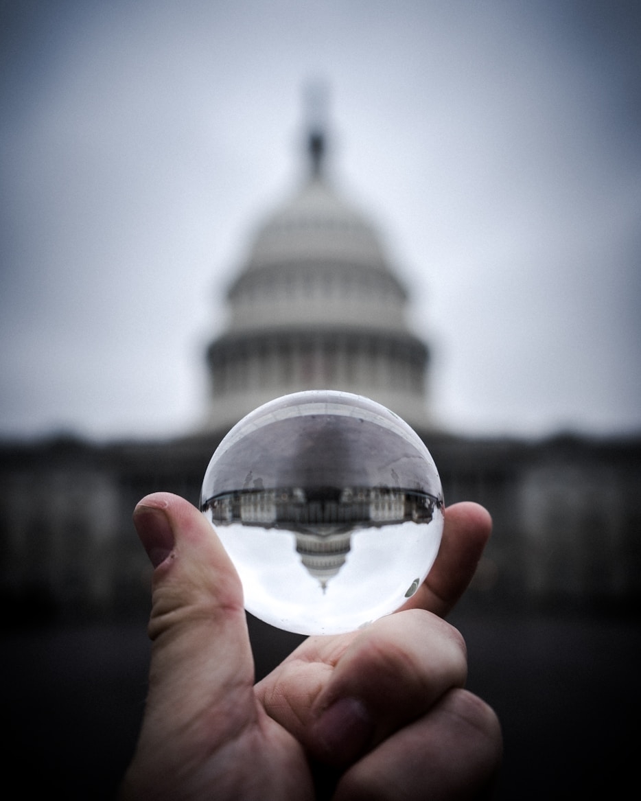 Una mano sostiene una bola de cristal que refleja el edificio del Capitolio en un fondo nublado.
