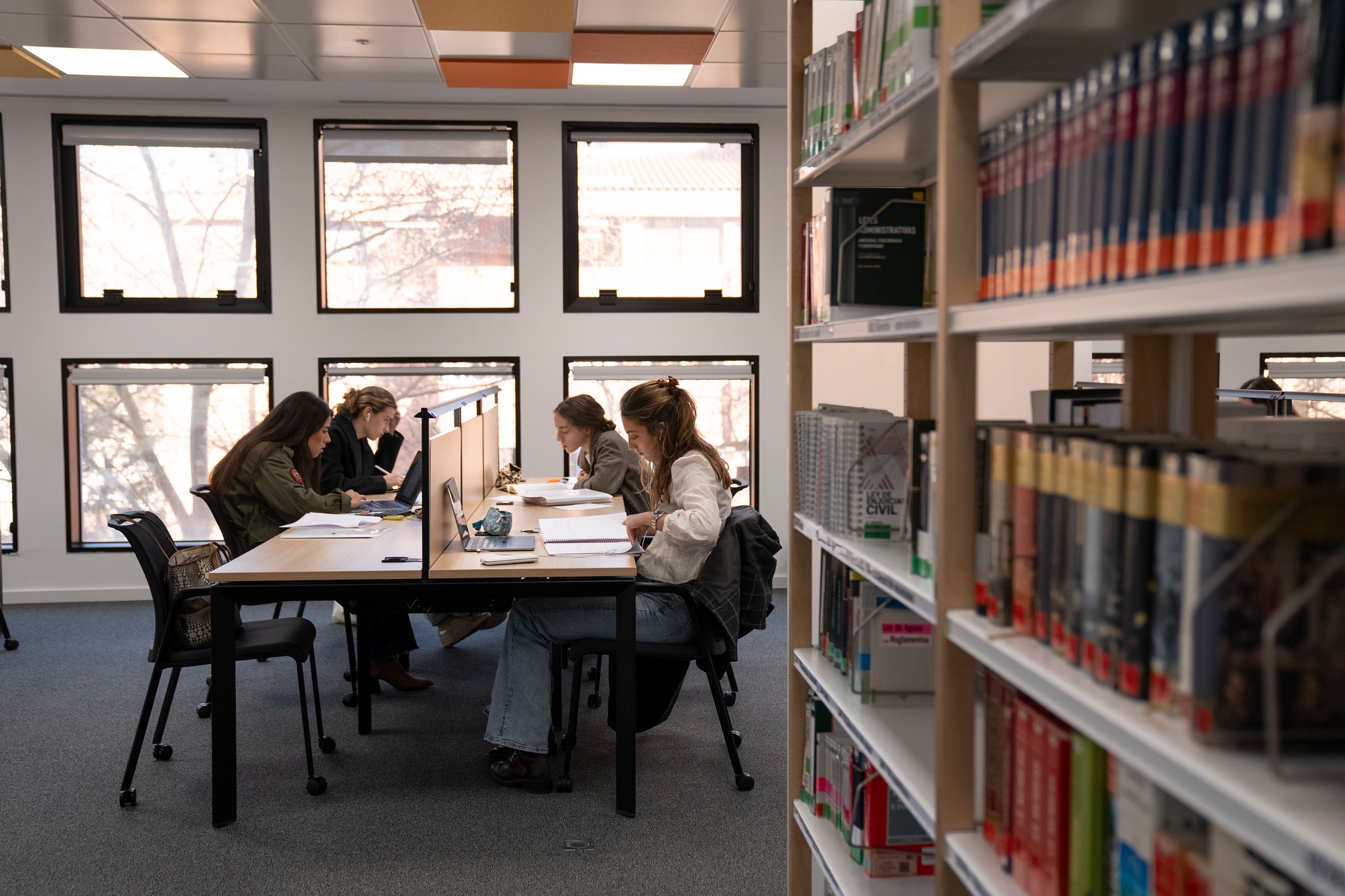 Un grupo de cuatro personas estudian en una biblioteca con mesas y estanterías de libros.