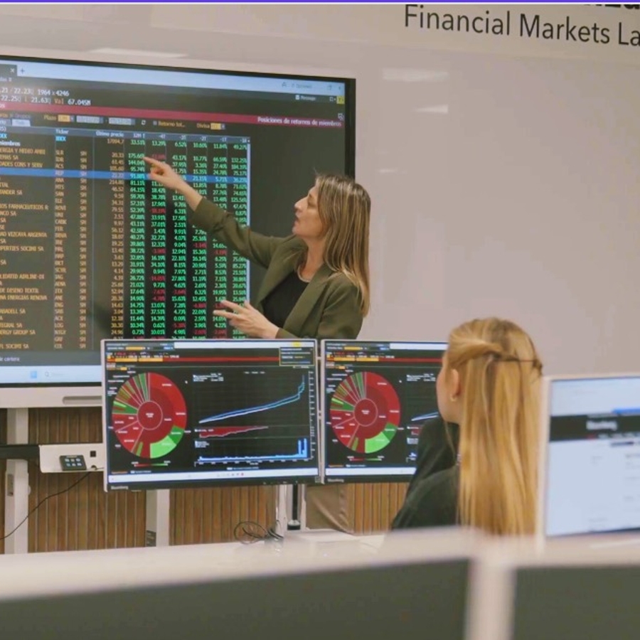 Una mujer está señalando gráficos en una pantalla grande mientras enseña a un grupo en un aula de finanzas.
