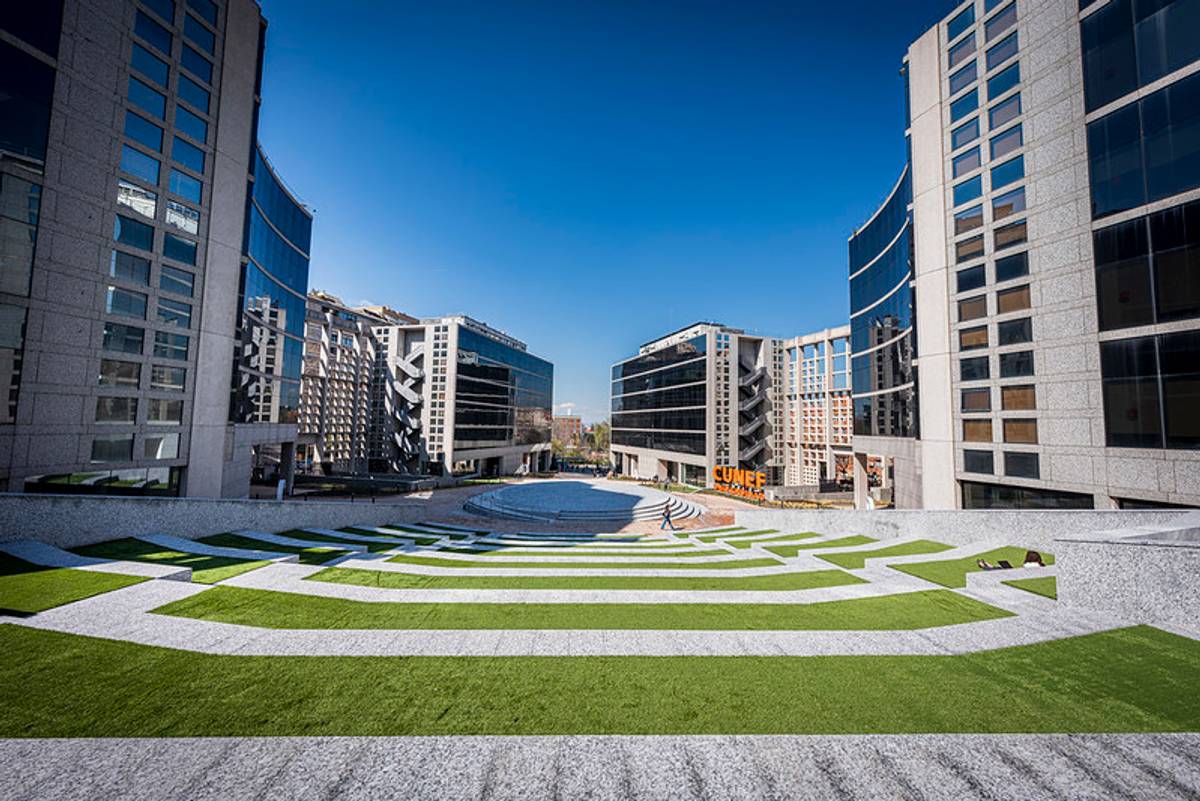 La imagen muestra un espacio urbano moderno con edificios de cristal y una gran área de césped en forma de espiral.