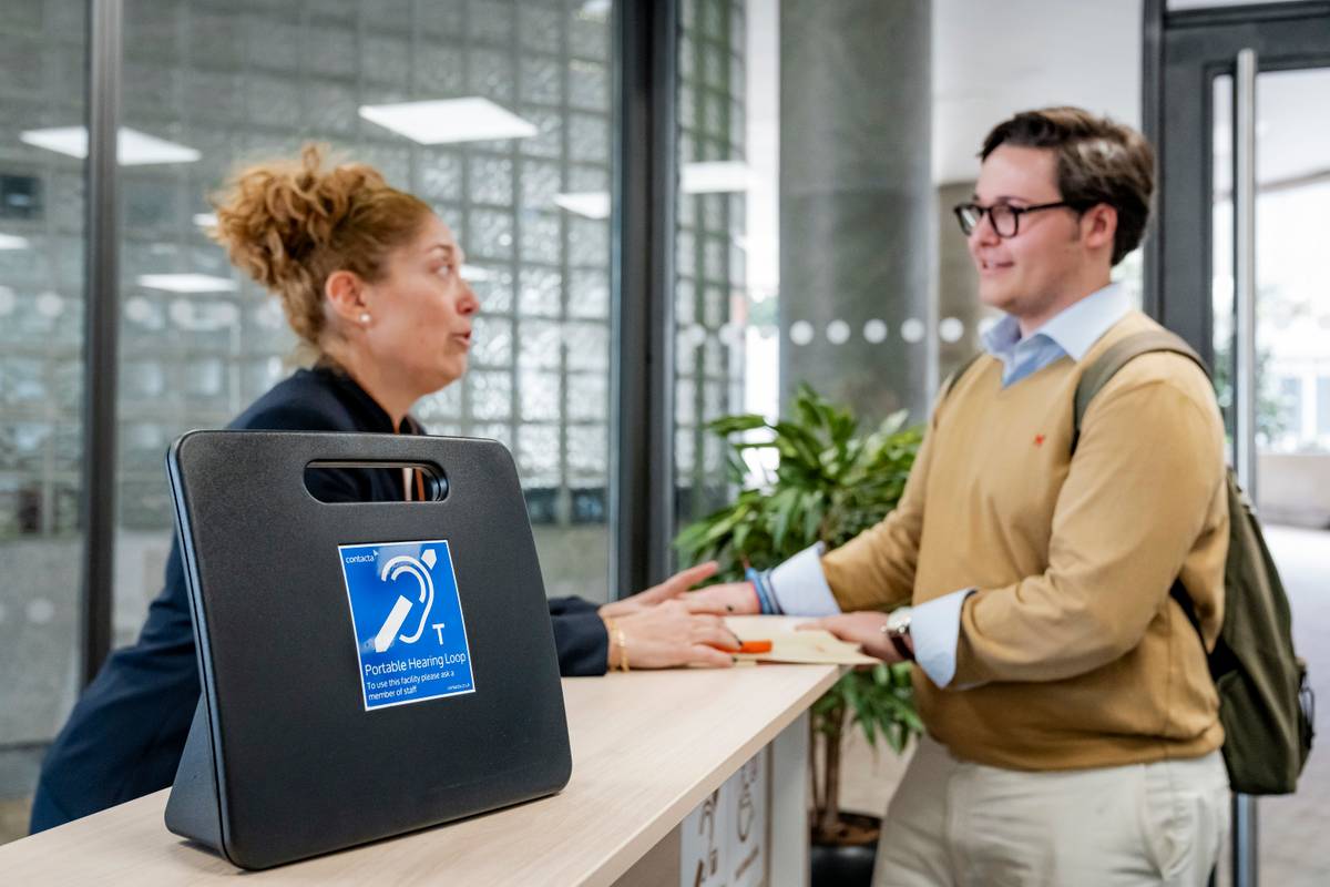 Una mujer conversa con un hombre en un mostrador, mientras un objeto con un símbolo de accesibilidad auditiva está en primer plano.