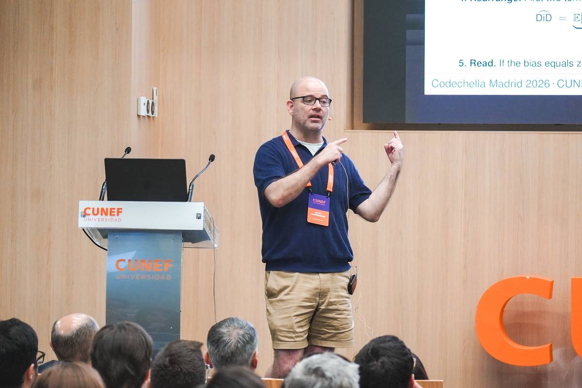Un ponente está hablando en una conferencia con una presentación en la pantalla detrás de él.