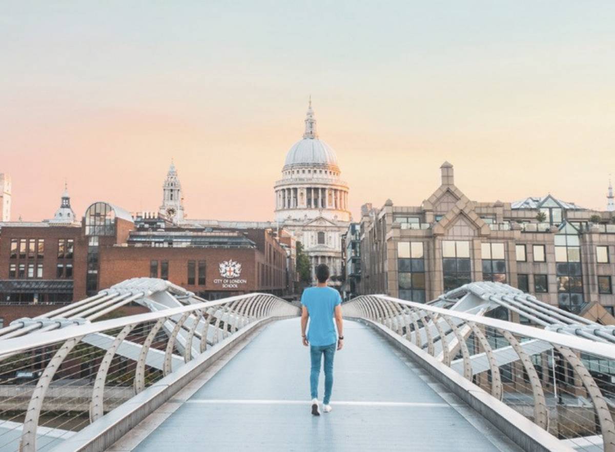 Una persona camina por un puente moderno con vistas a una catedral y edificios urbanos al atardecer.