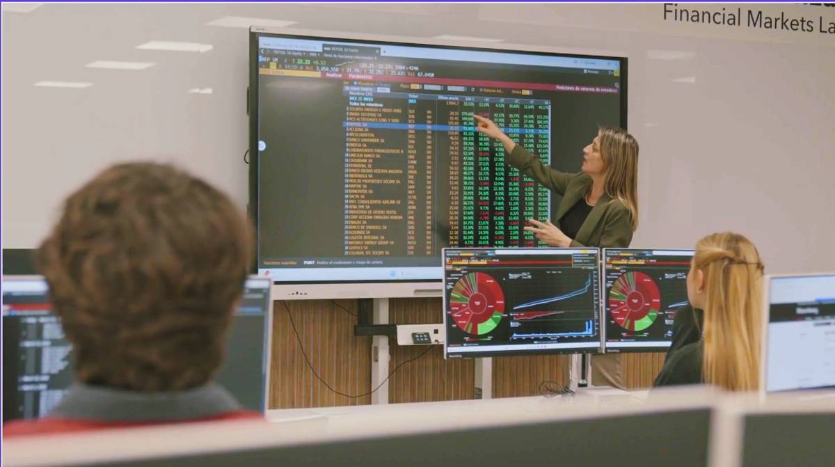 Una mujer presenta datos en una pantalla en una sala de clases sobre mercados financieros.