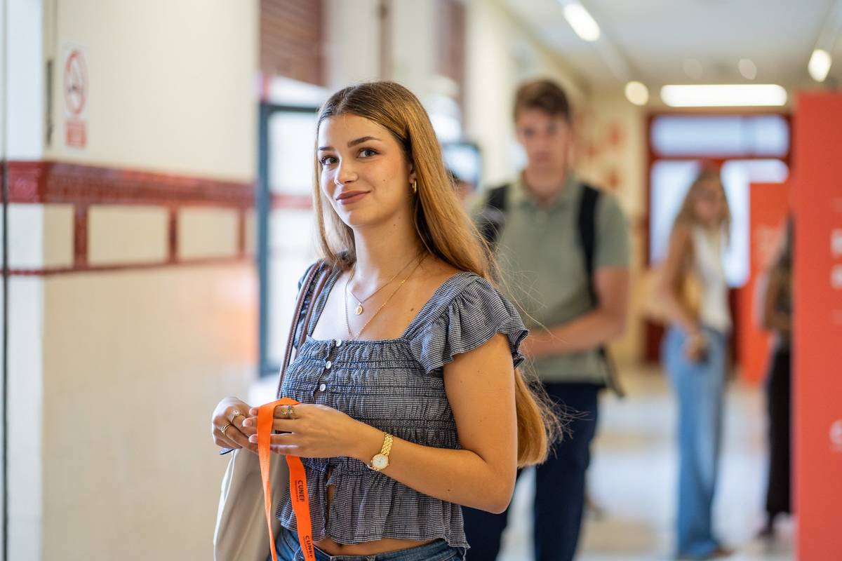Una joven sonriente se encuentra en un pasillo con otras personas al fondo.
