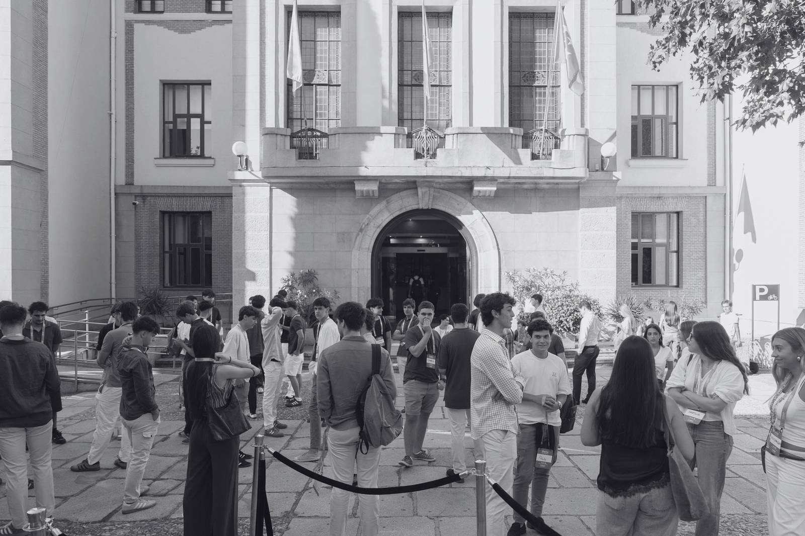Un grupo de personas socializa frente a un edificio con banderas en un ambiente exterior.