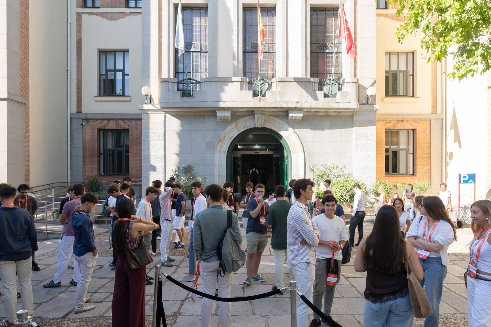 Un grupo de estudiantes se reúne en la entrada de un edificio con banderas y un ambiente animado.