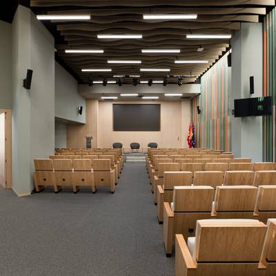 Sala moderna y luminosa destinada a conferencias, con asientos de madera y un gran panel de presentación.