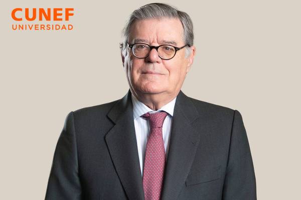 Retrato de un hombre con gafas, vestido con un traje oscuro y corbata, frente a un fondo beige.