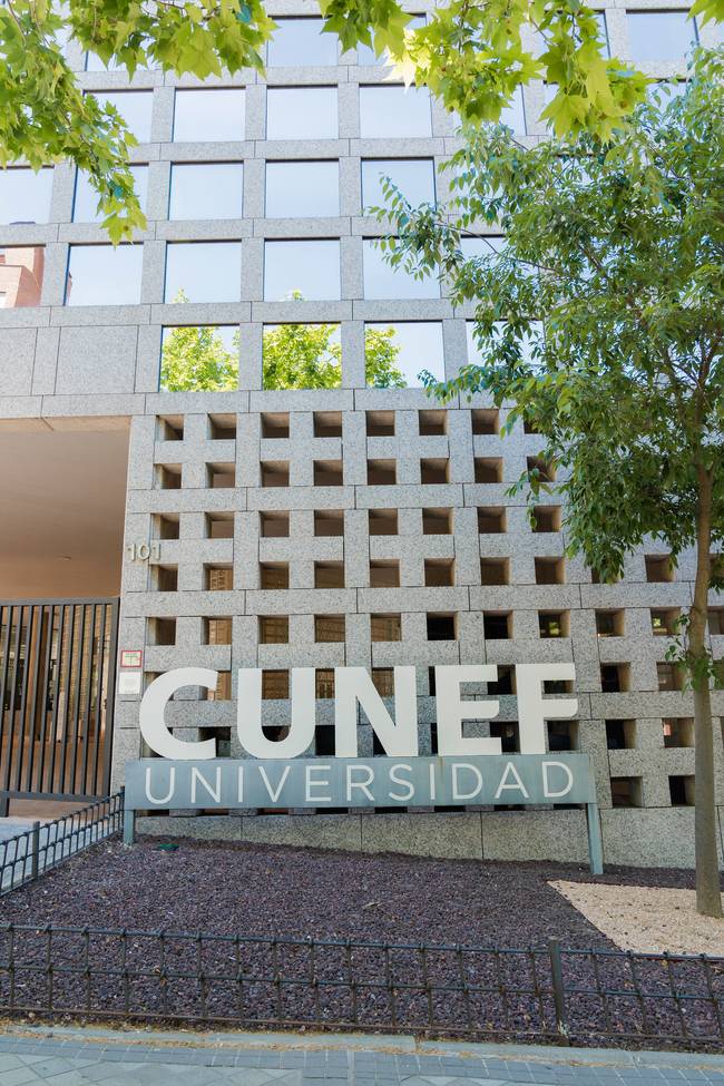 La imagen muestra la entrada de la Universidad CUNEF con un diseño arquitectónico moderno y árboles alrededor.