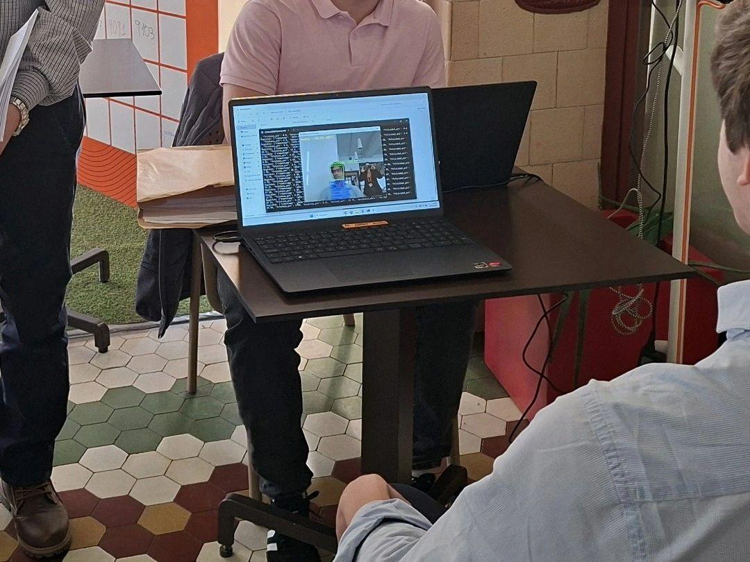 Una reunión virtual en la que se ve a una persona en la pantalla de una laptop mientras dos personas están sentadas alrededor de una mesa.