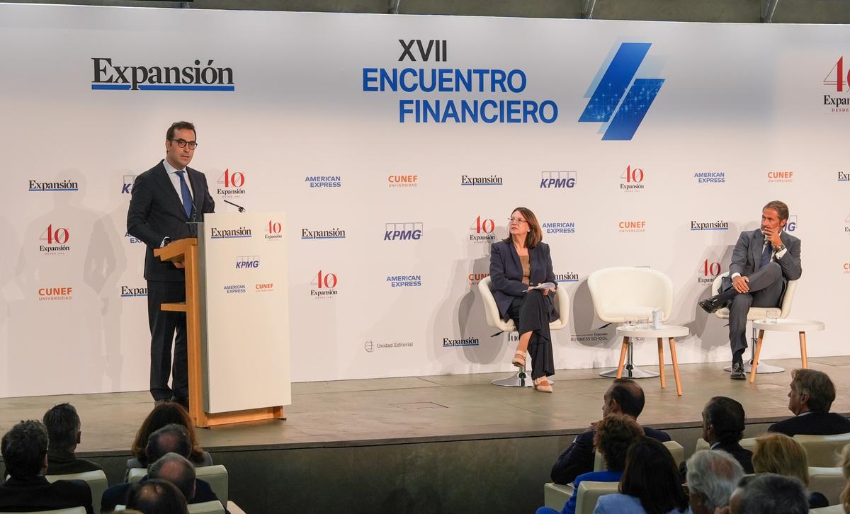 Un evento de encuentro financiero con un ponente en el podio y dos participantes sentados ante una audiencia.