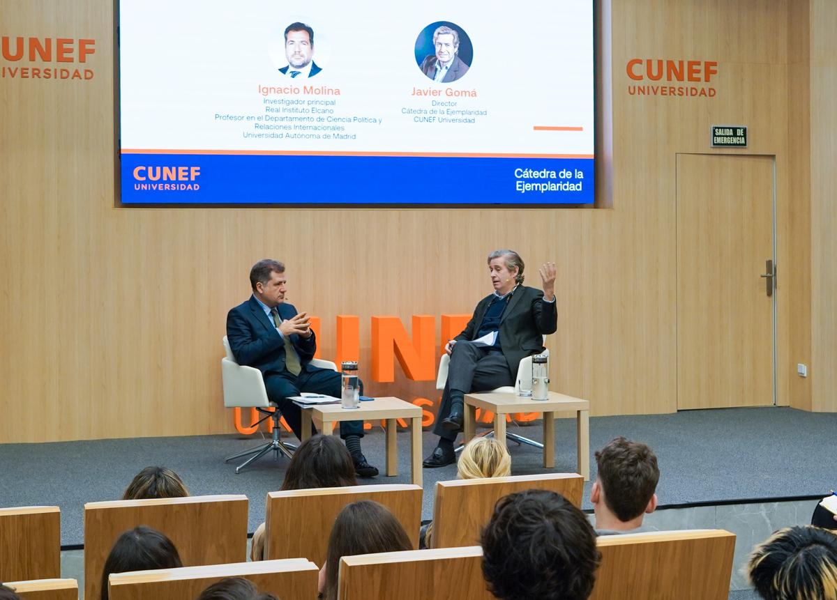 Dos hombres discuten en un evento académico en CUNEF, mientras un público los observa atentamente.