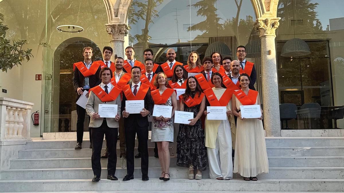 Un grupo de graduados posando en la entrada de un edificio con trajes formales y togas naranjas.