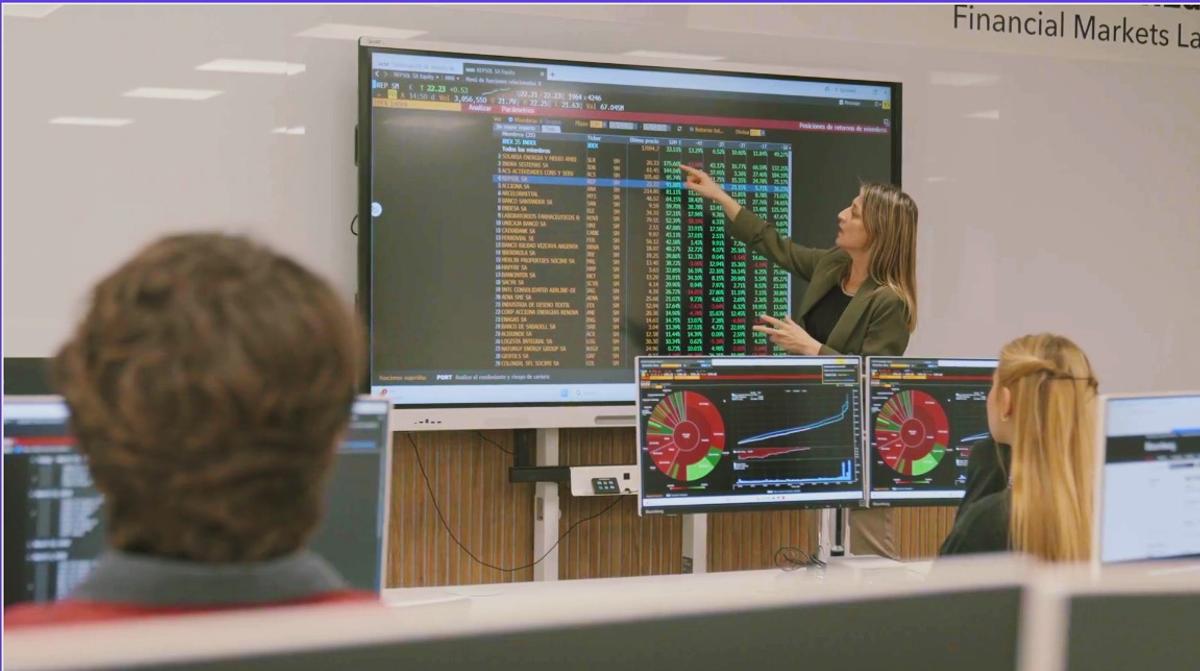 Una mujer está señalando un gráfico en una pantalla grande mientras otras personas la observan en un entorno de aula de finanzas.