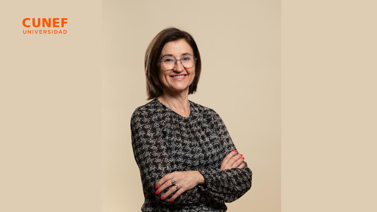 Una mujer de pie con los brazos cruzados y una sonrisa, viste una blusa de patrón oscuro y tiene el cabello corto.