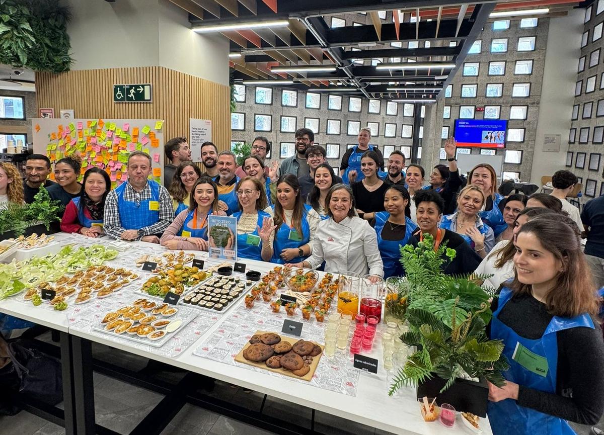 Un grupo de personas sonrientes se reúne alrededor de una mesa llena de diversos platillos y bebidas en un evento comunitario.