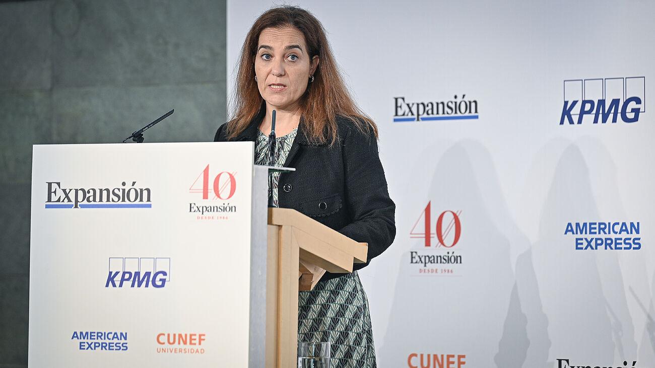Una mujer hablando en un evento profesional frente a un estrado con logotipos de varias empresas.
