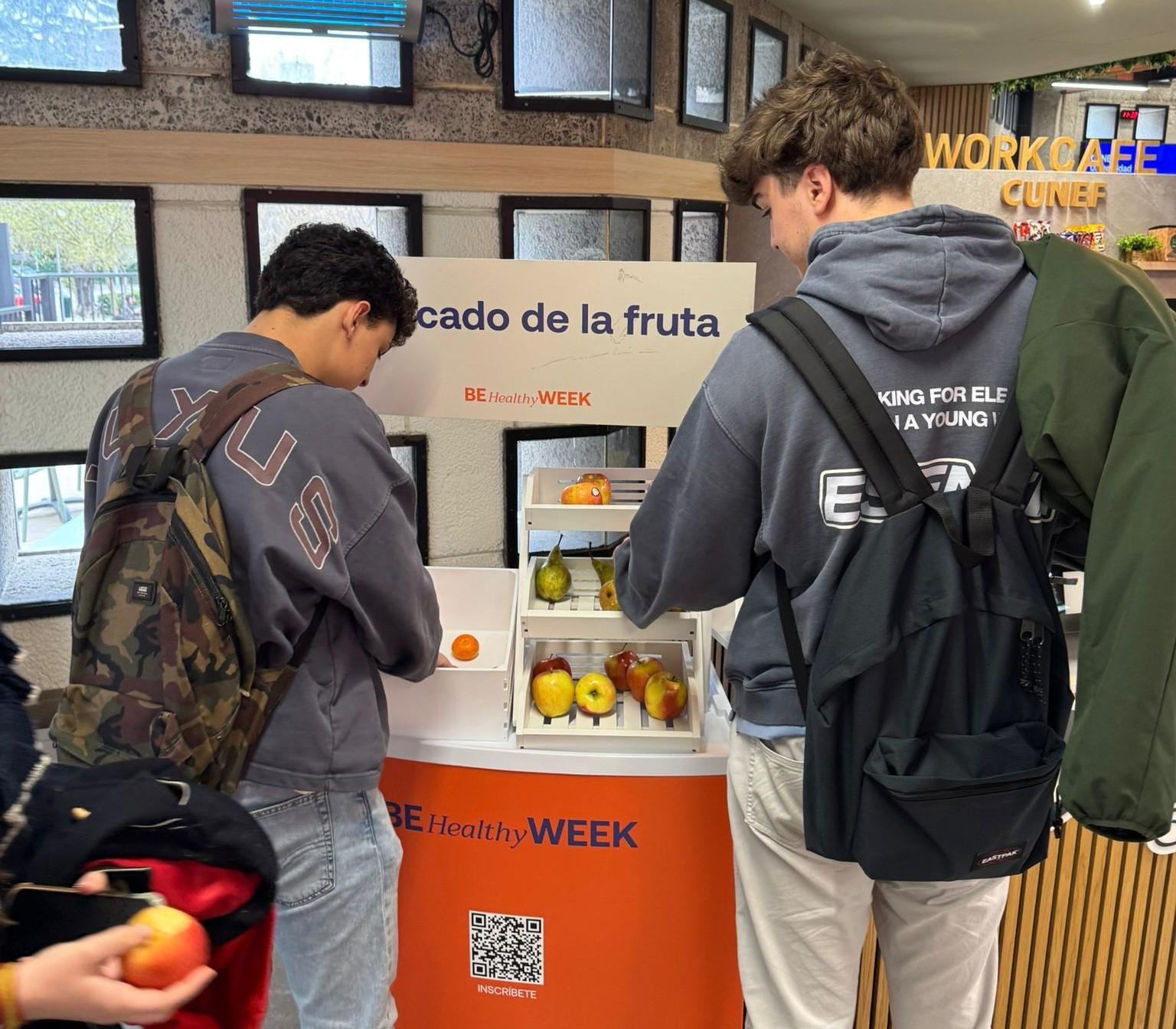 Dos jóvenes interactúan con un dispensador de frutas en un evento de promoción de la salud.
