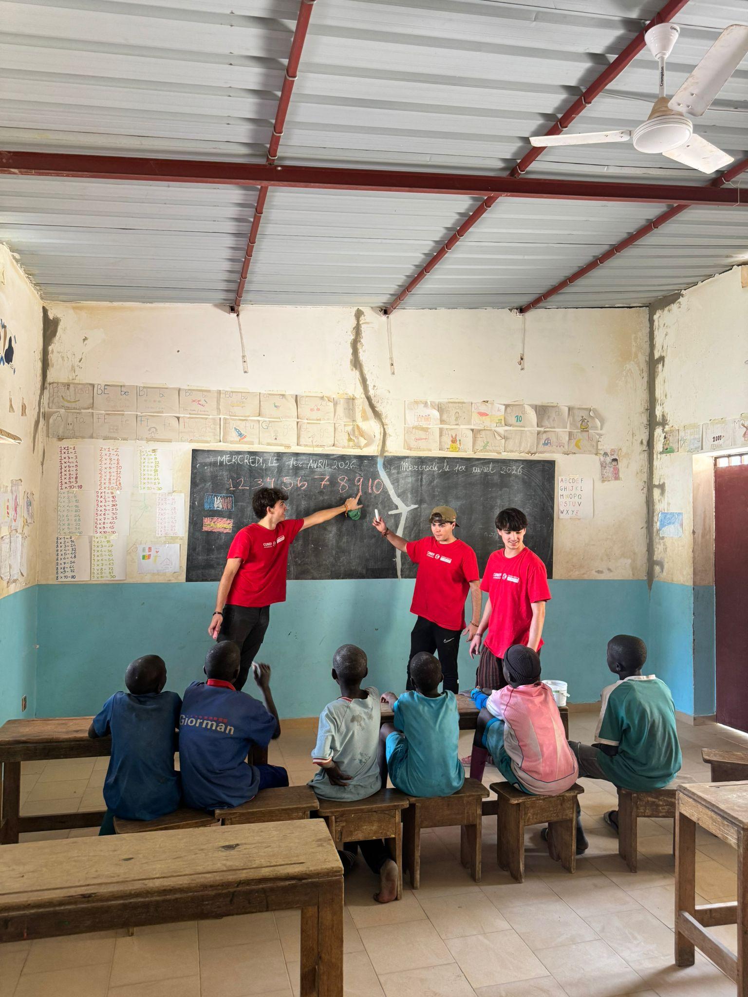 Un grupo de jóvenes enseña en un aula a un grupo de niños sentados en bancos de madera.