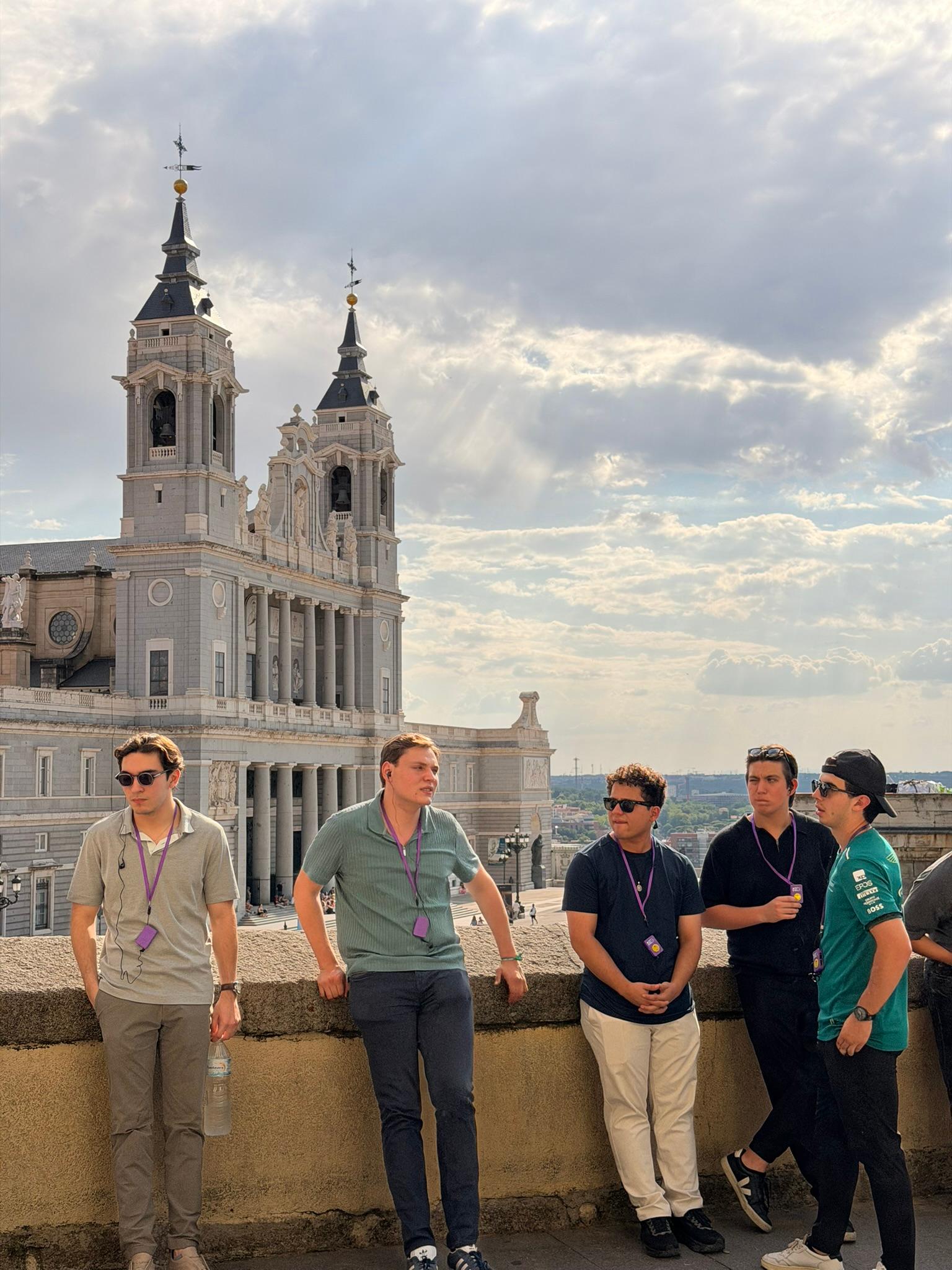 Un grupo de hombres conversando frente a un edificio monumental con torres, bajo un cielo nublado.