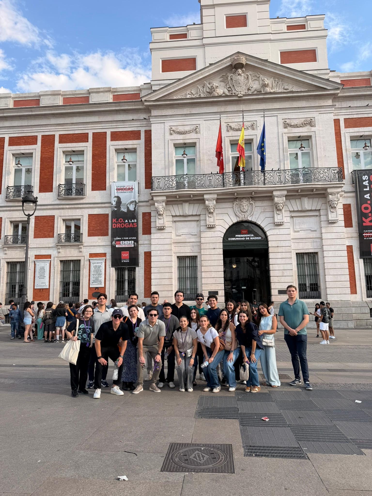 Un grupo de personas posa frente a un edificio emblemático en una plaza con cielo despejado.