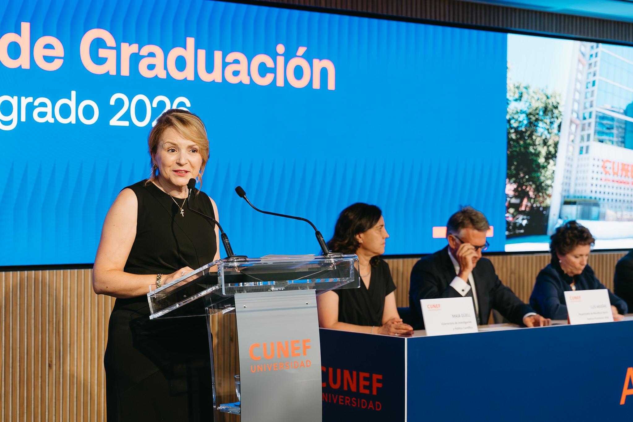 Una mujer habla desde un podio durante una ceremonia de graduación con un panel de asistentes en el fondo.