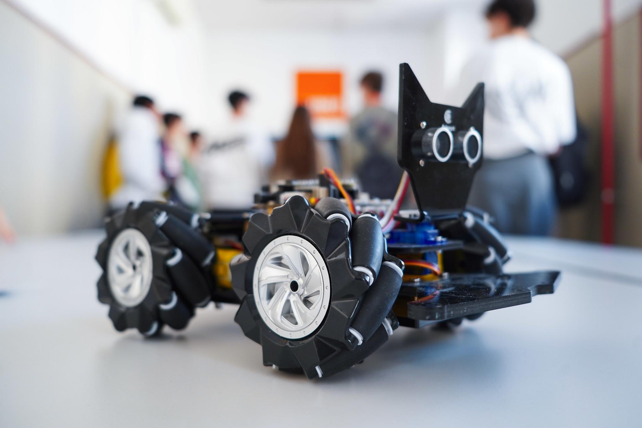 Un robot de cuatro ruedas diseñado con un cuerpo negro y detalles en plata, se encuentra en primer plano sobre una mesa mientras al fondo hay personas en conversación.