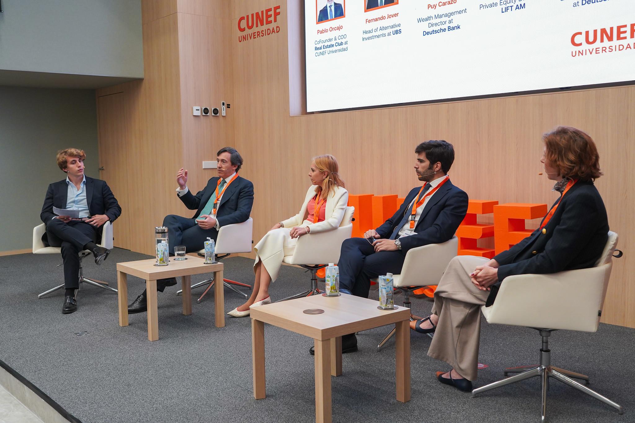 Un panel de discusión con cinco ponentes sentados, presentando sus ideas en un auditorio moderno.
