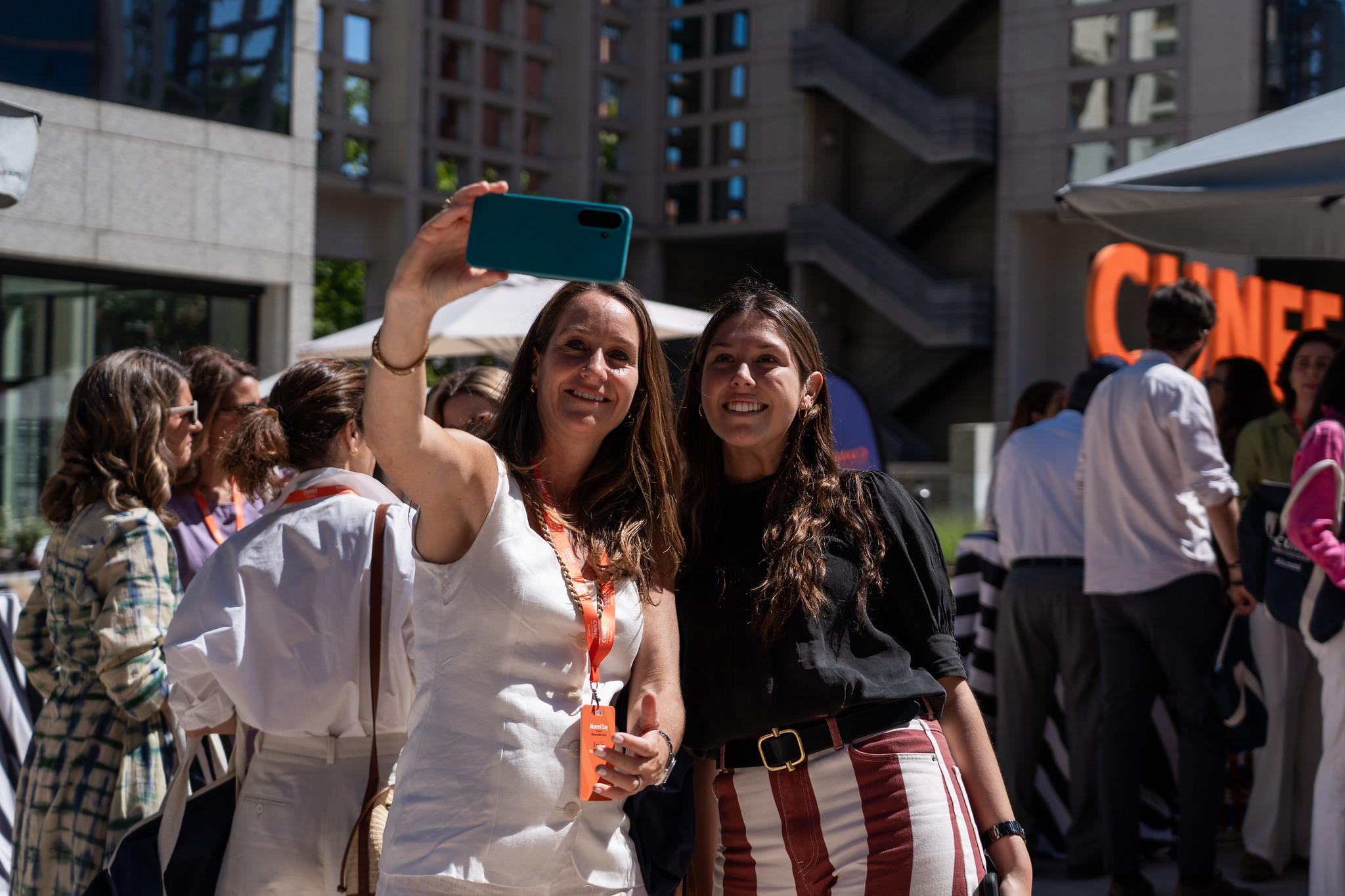 Dos mujeres se toman una selfie en un evento al aire libre con fondo de personas y edificios.