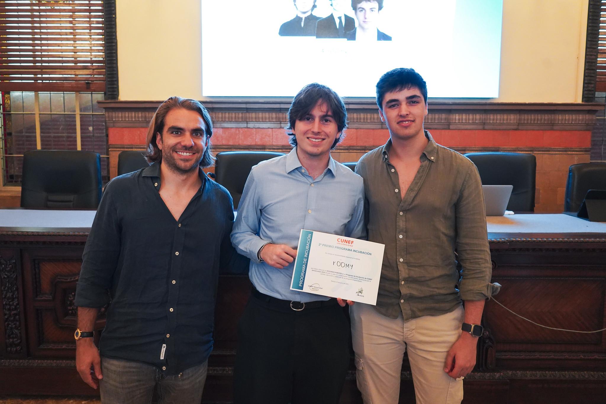 Tres jóvenes posan sonrientes sosteniendo un certificado en un evento.