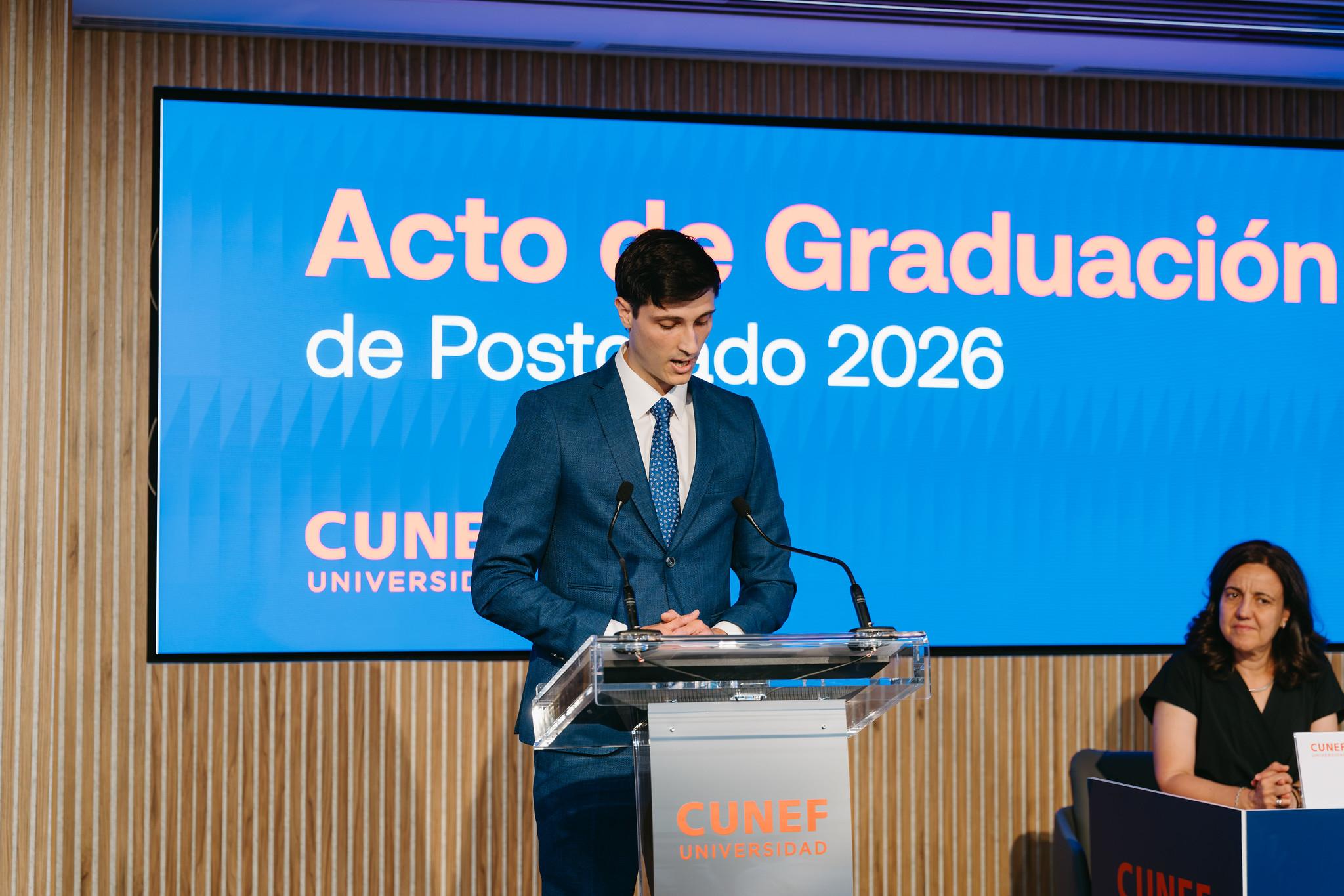Un hombre joven, vestido de traje, habla en un podium durante la ceremonia de graduación de un programa de posgrado.