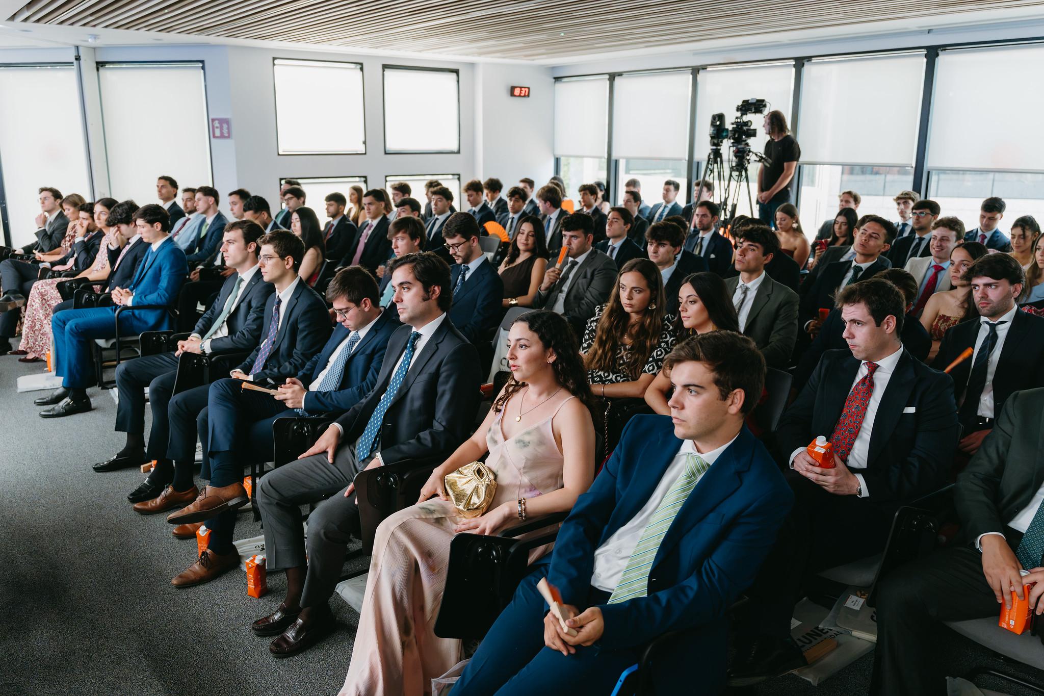 Una audiencia numerosa sentada en un salón, atenta a una presentación.