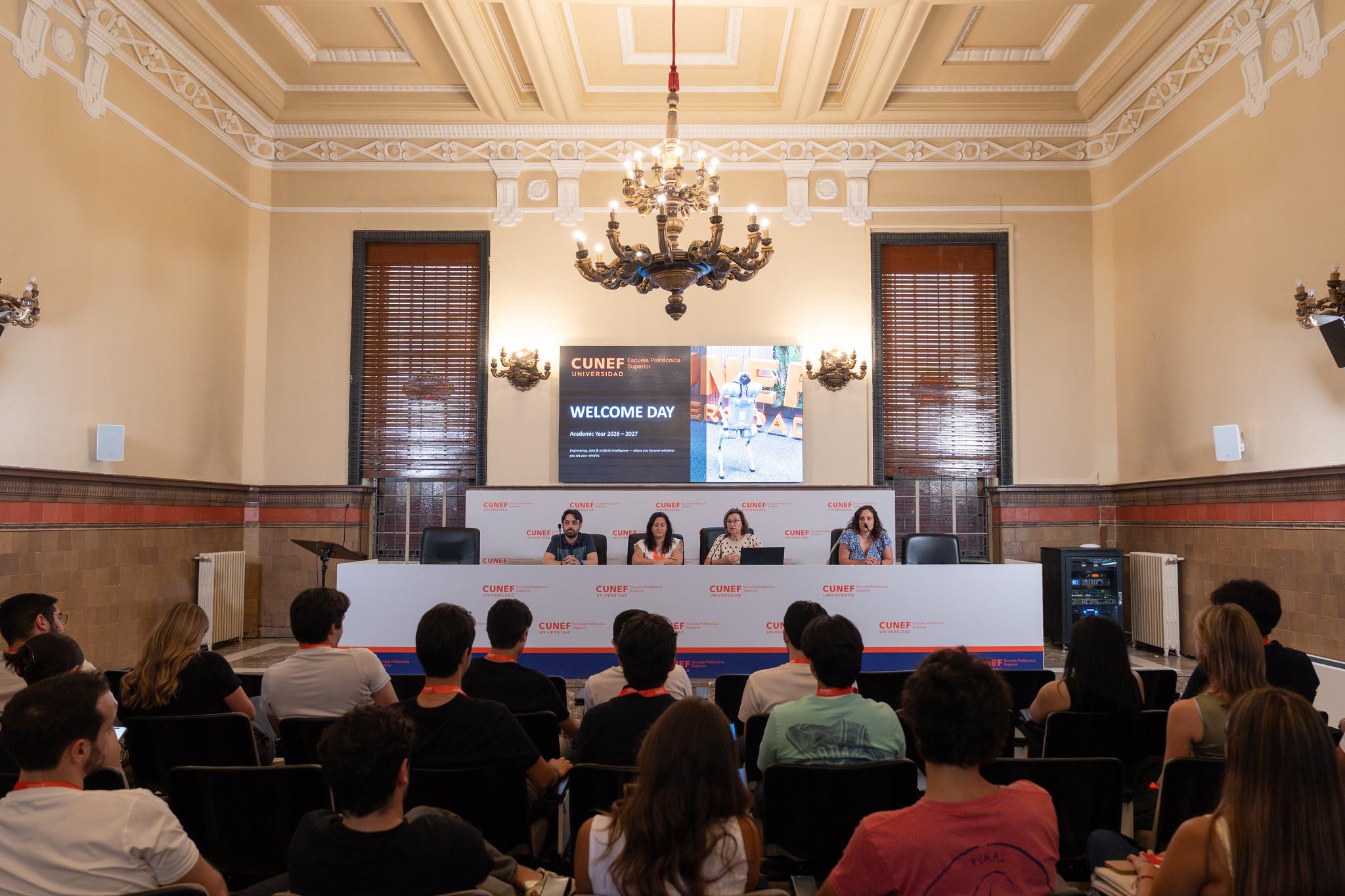 Un grupo de personas asiste a un evento de bienvenida en una sala formal con panelistas hablando en un escenario.