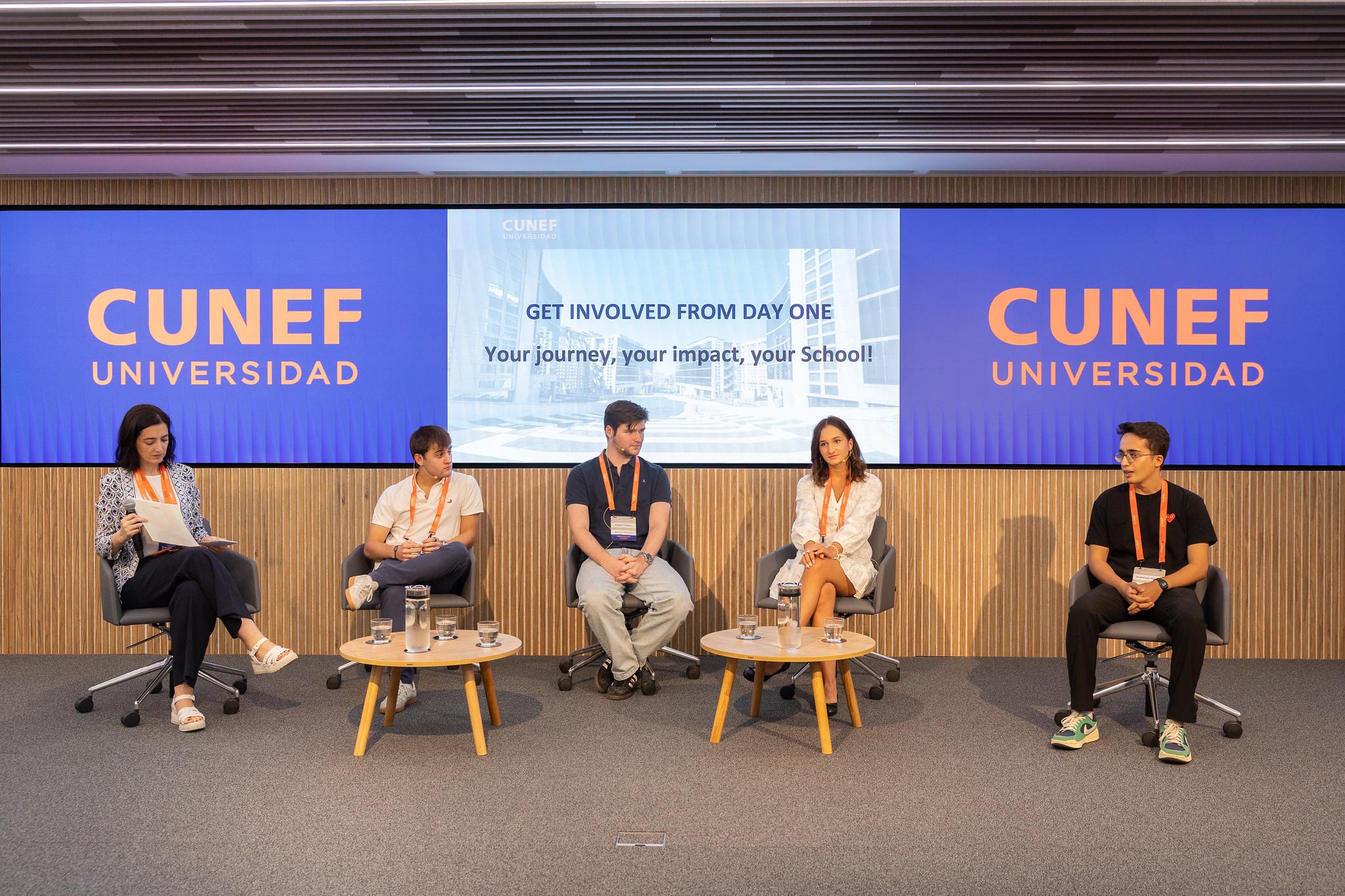 Imagen de un panel de discusión en una universidad con cuatro participantes sentados frente a una audiencia.