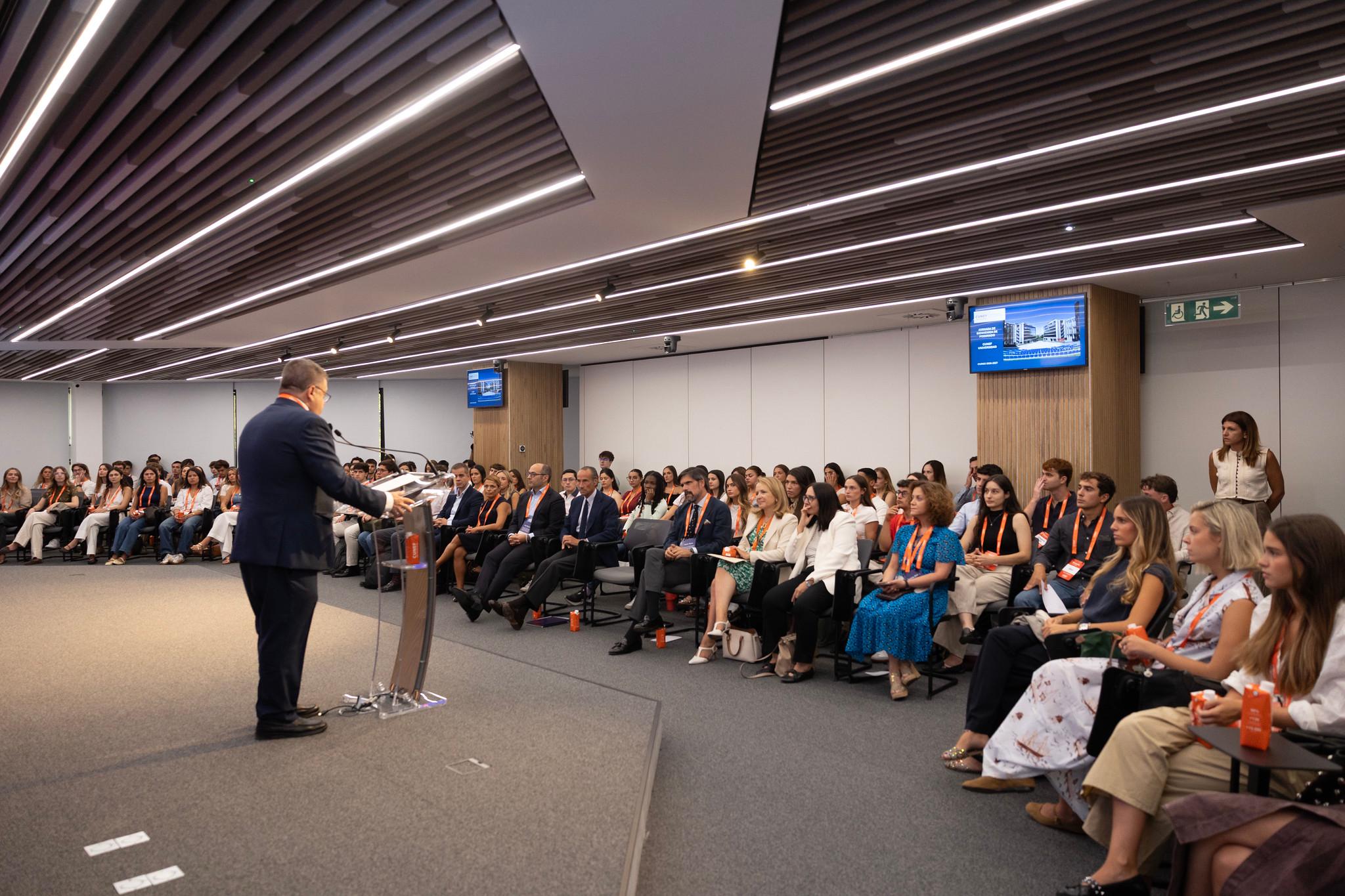 Un ponente habla frente a un público numeroso en un evento en una sala moderna.