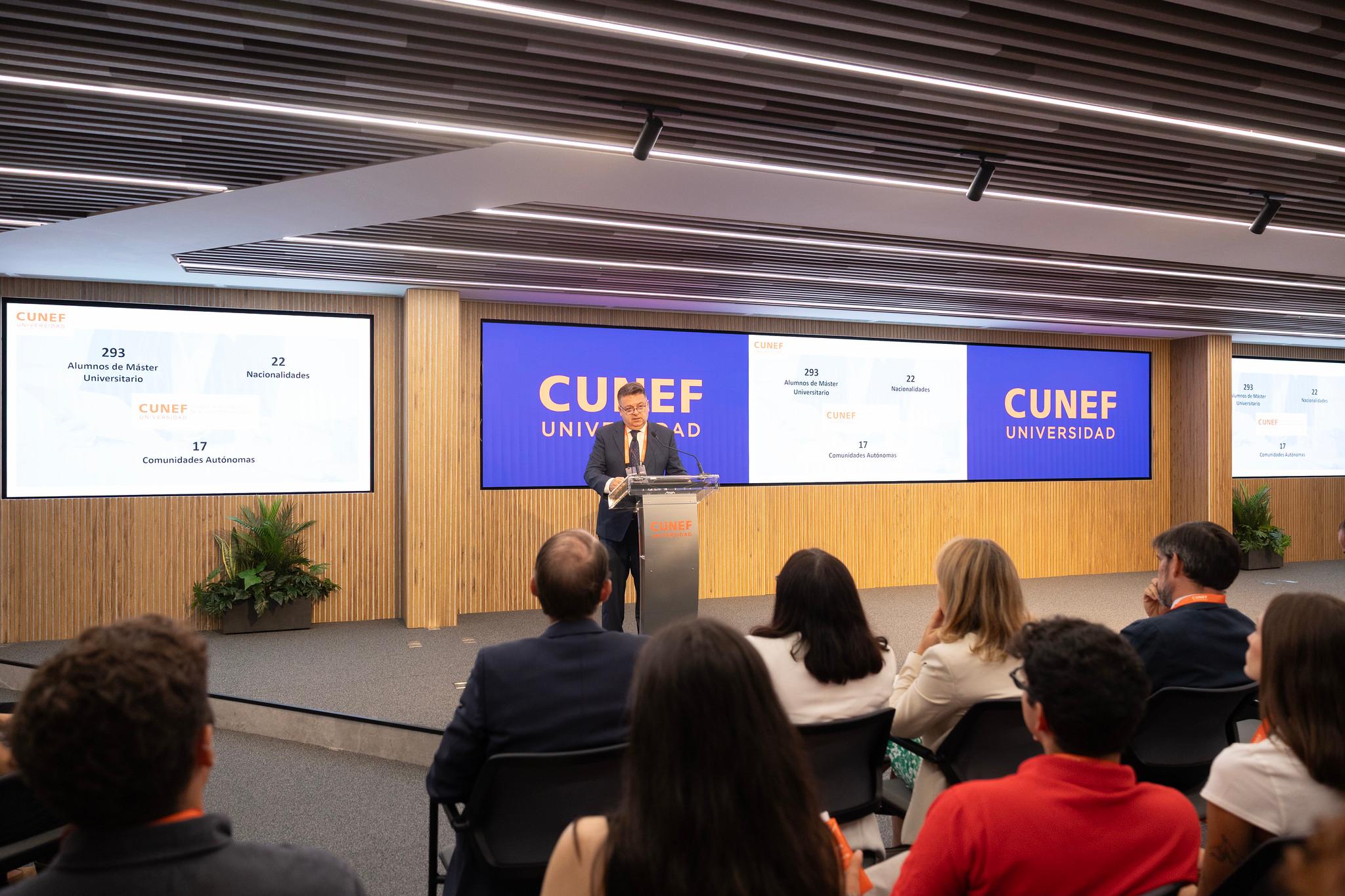 Un presentador habla en un auditorio de la Universidad CUNEF mientras una presentación se muestra en pantallas grandes.