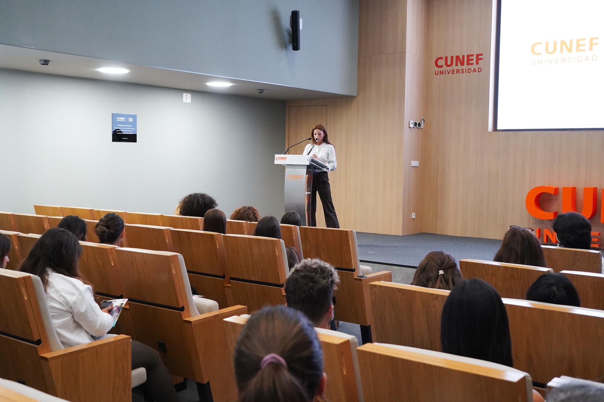Una mujer habla frente a un auditorio en una universidad mientras los asistentes la escuchan.