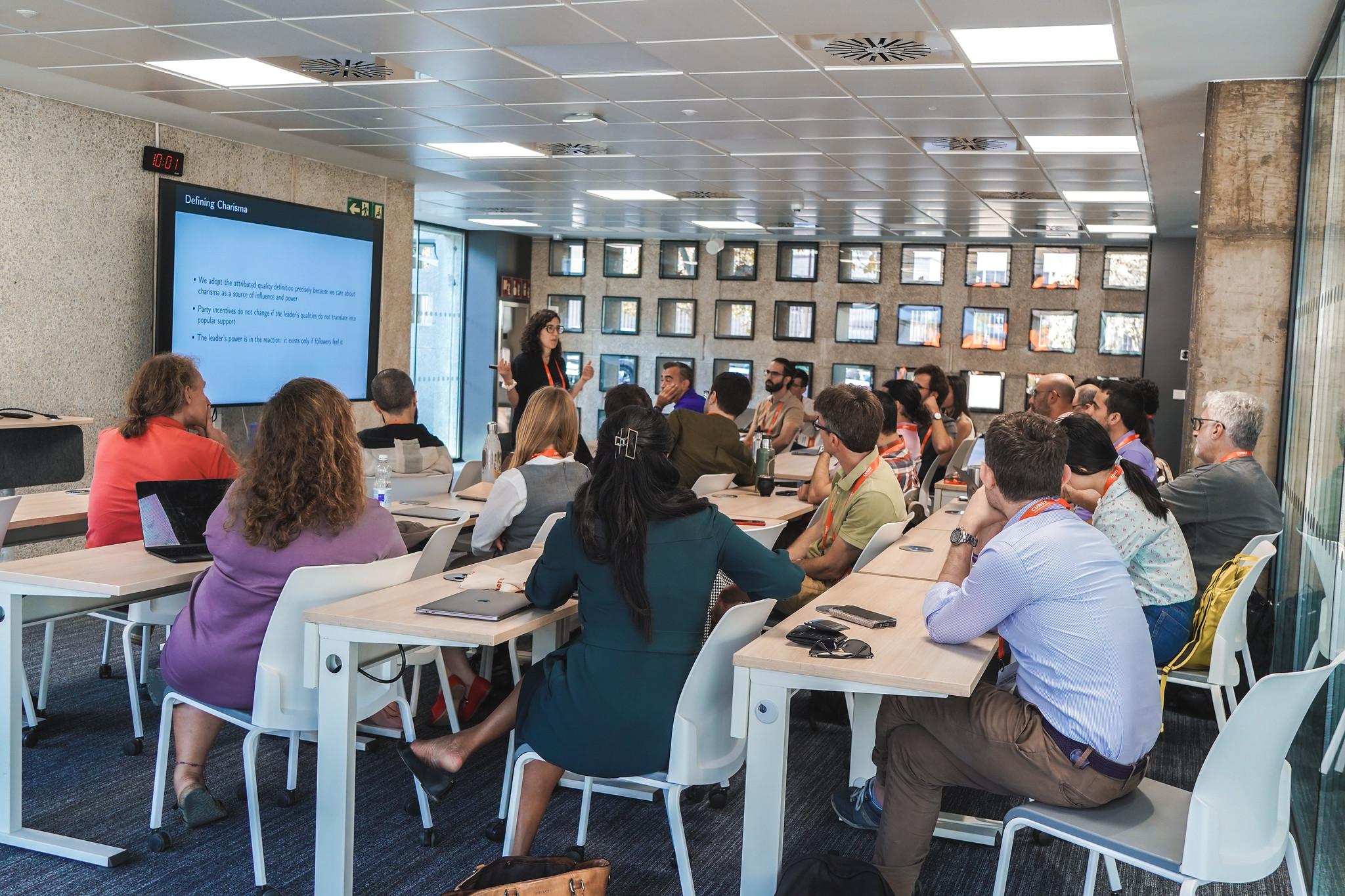 La imagen muestra una reunión en una sala con un grupo de personas sentadas y una presentadora frente a una pantalla.