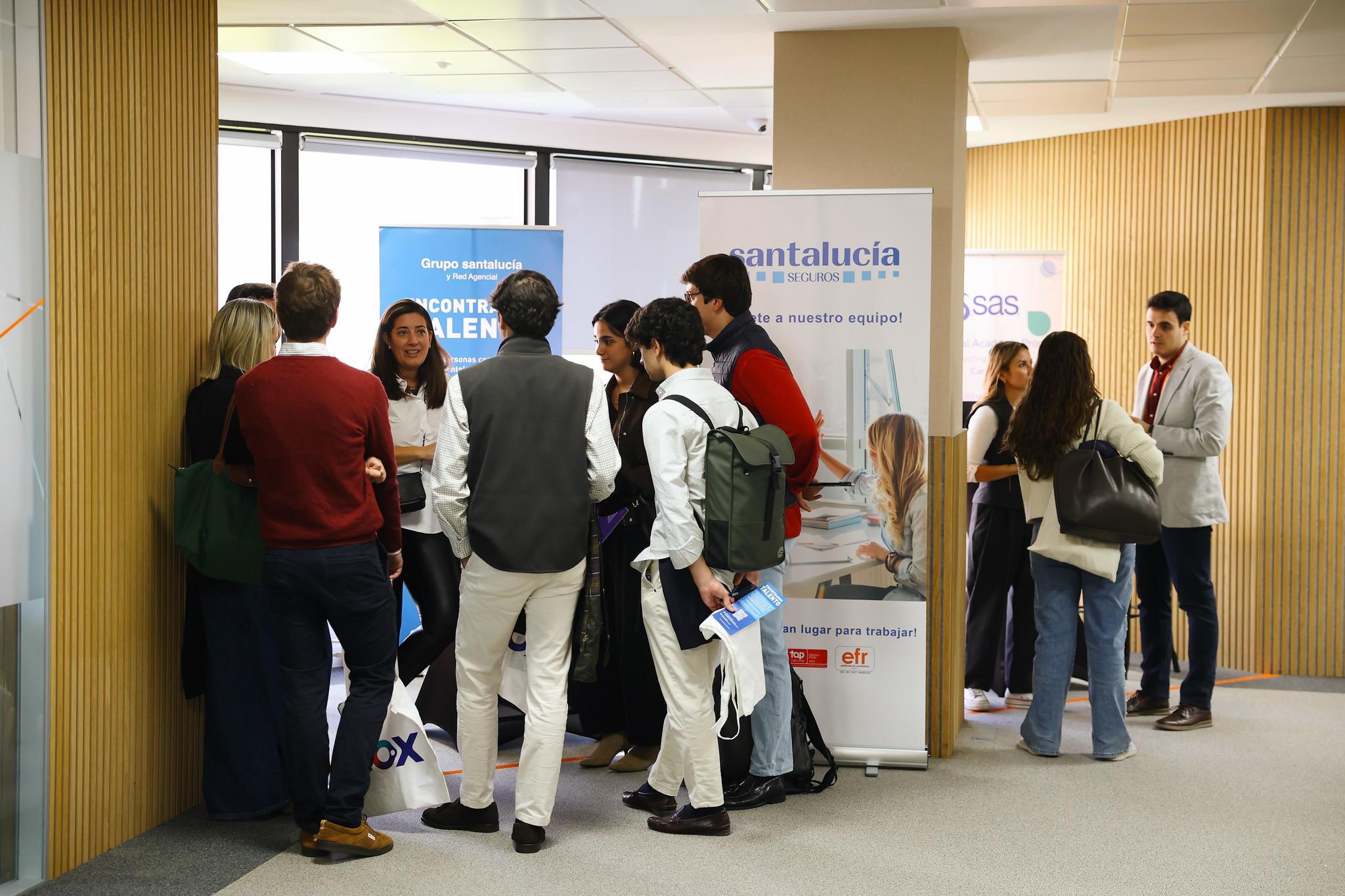 Un grupo de personas interactúa en un evento profesional en una sala con diferentes stands.