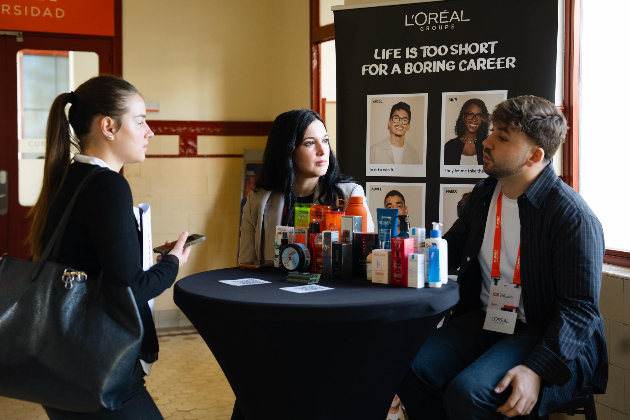 Una conversación entre tres personas frente a un stand de L'Oréal en un evento profesional.