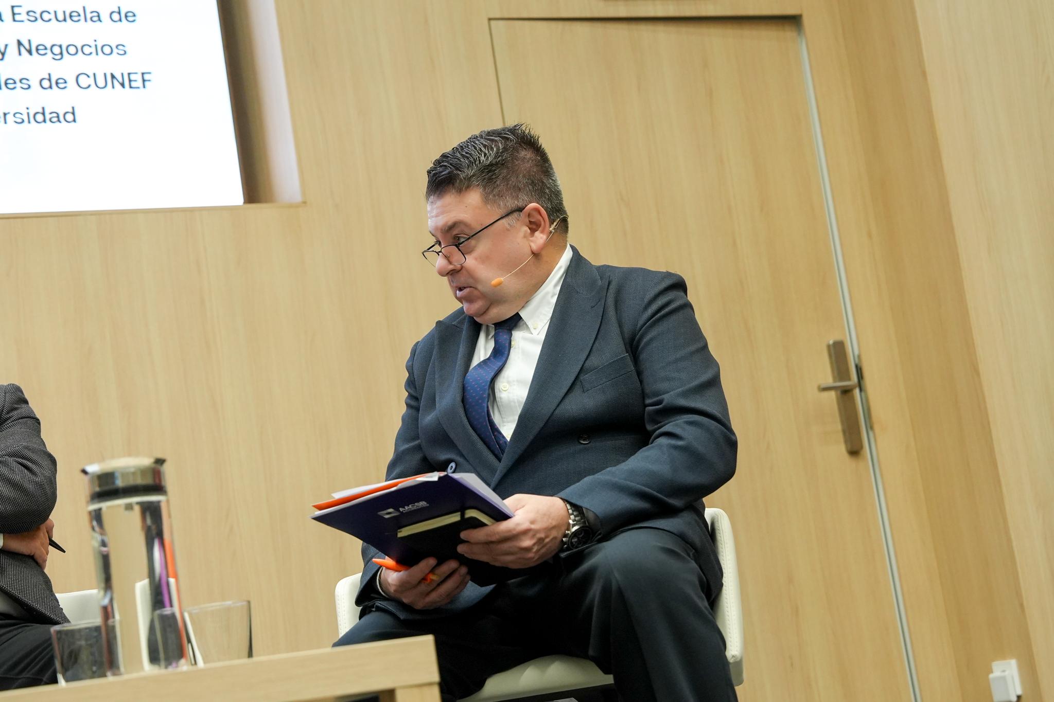 Un hombre vestido formalmente sentado y sosteniendo un libro en un evento en un entorno académico.