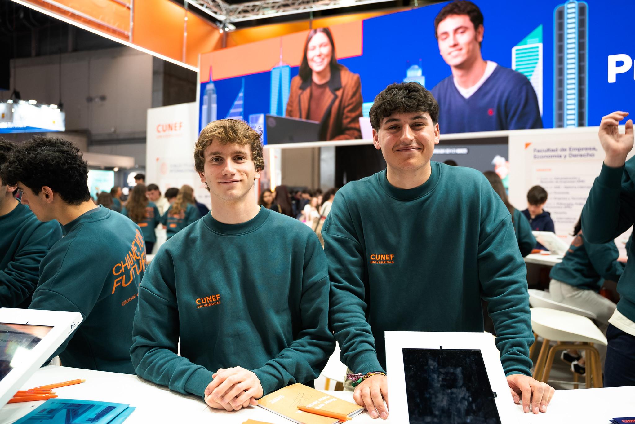 Dos jóvenes con sudaderas verdes sonríen en un evento, rodeados de pantallas y mesas de trabajo.