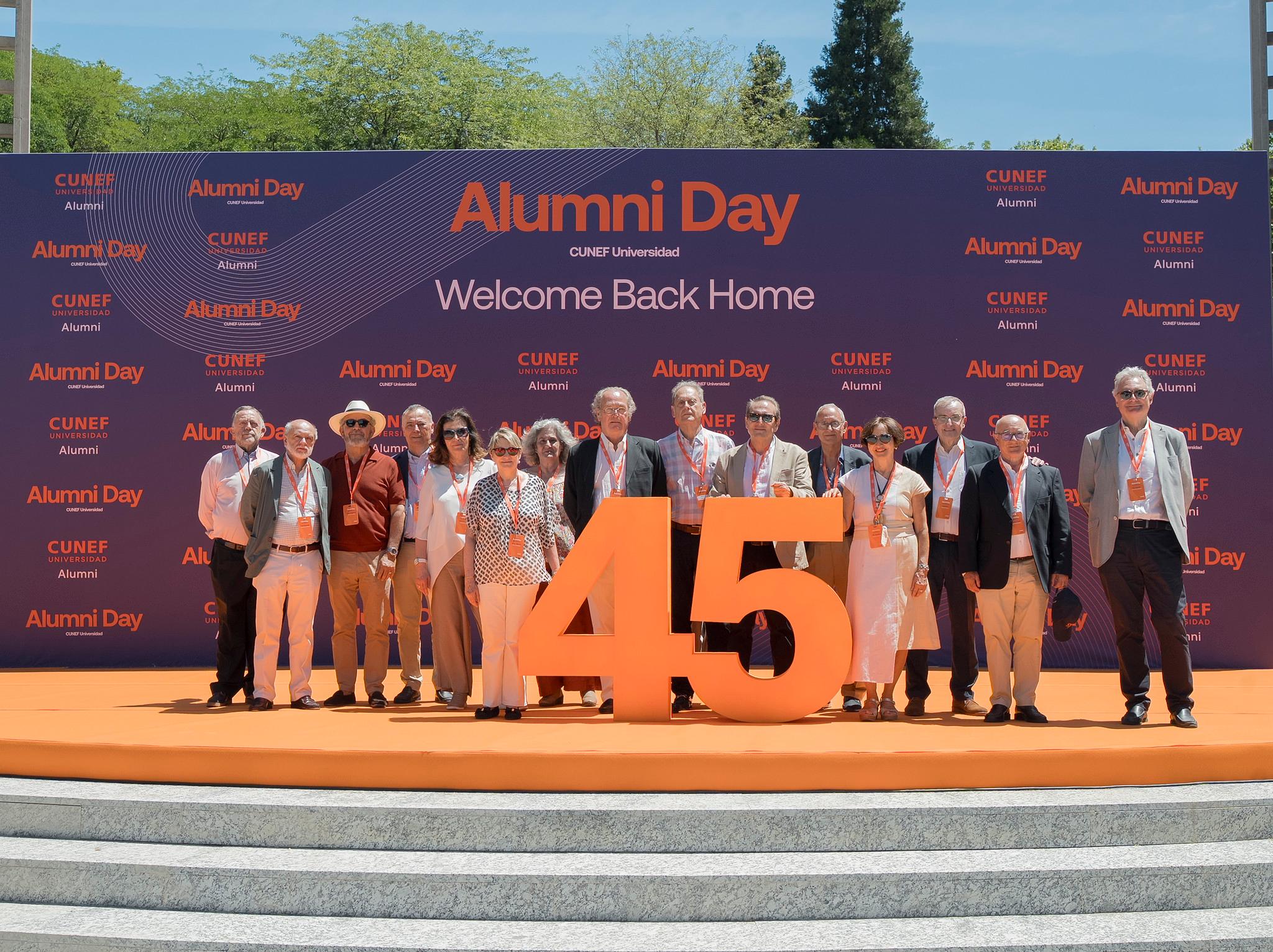 Un grupo de personas celebra el Alumni Day en un evento al aire libre con un gran número 45 en el centro.