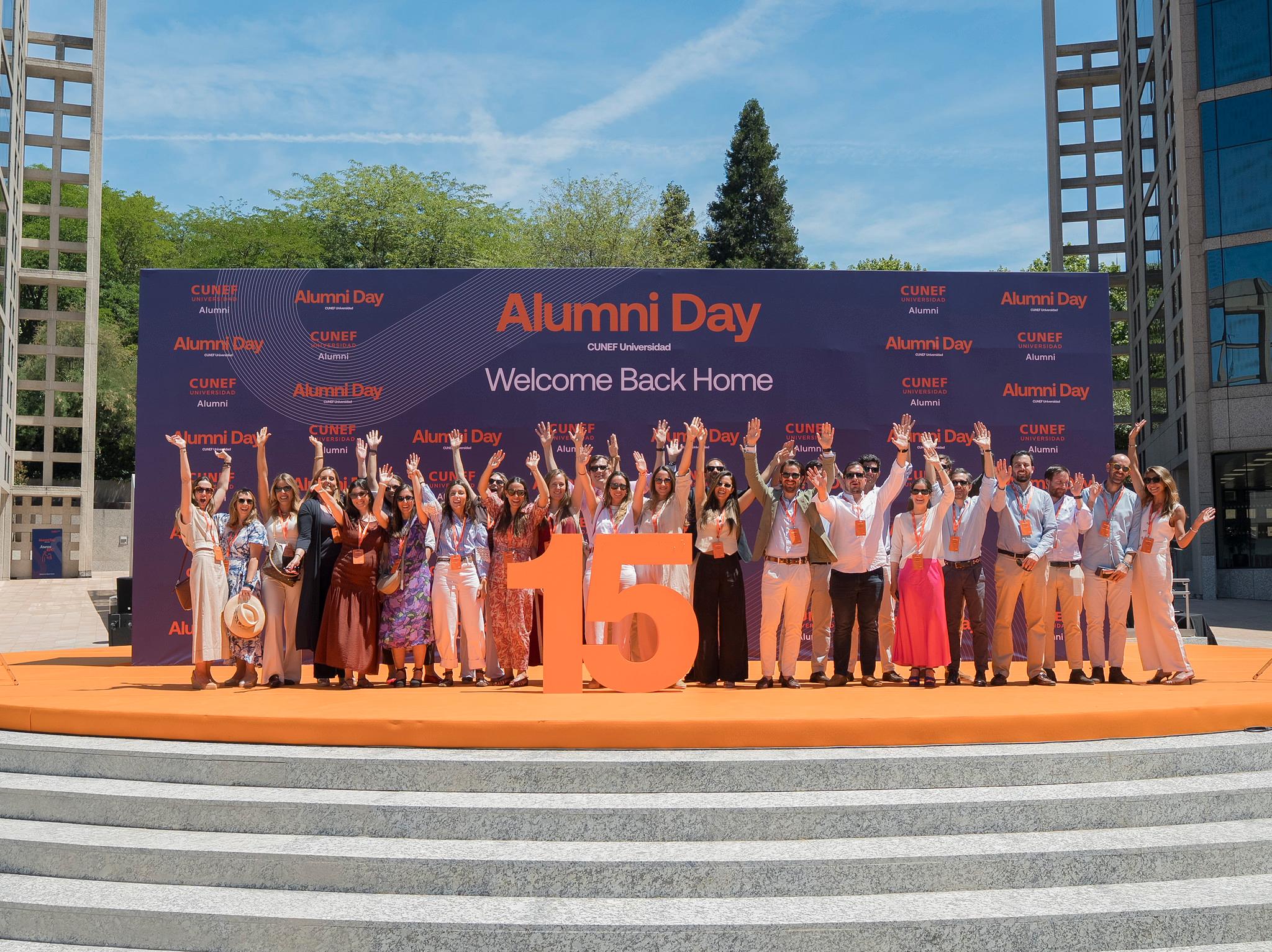 Un grupo de personas celebra el Alumni Day en un evento al aire libre con una gran pancarta detrás.