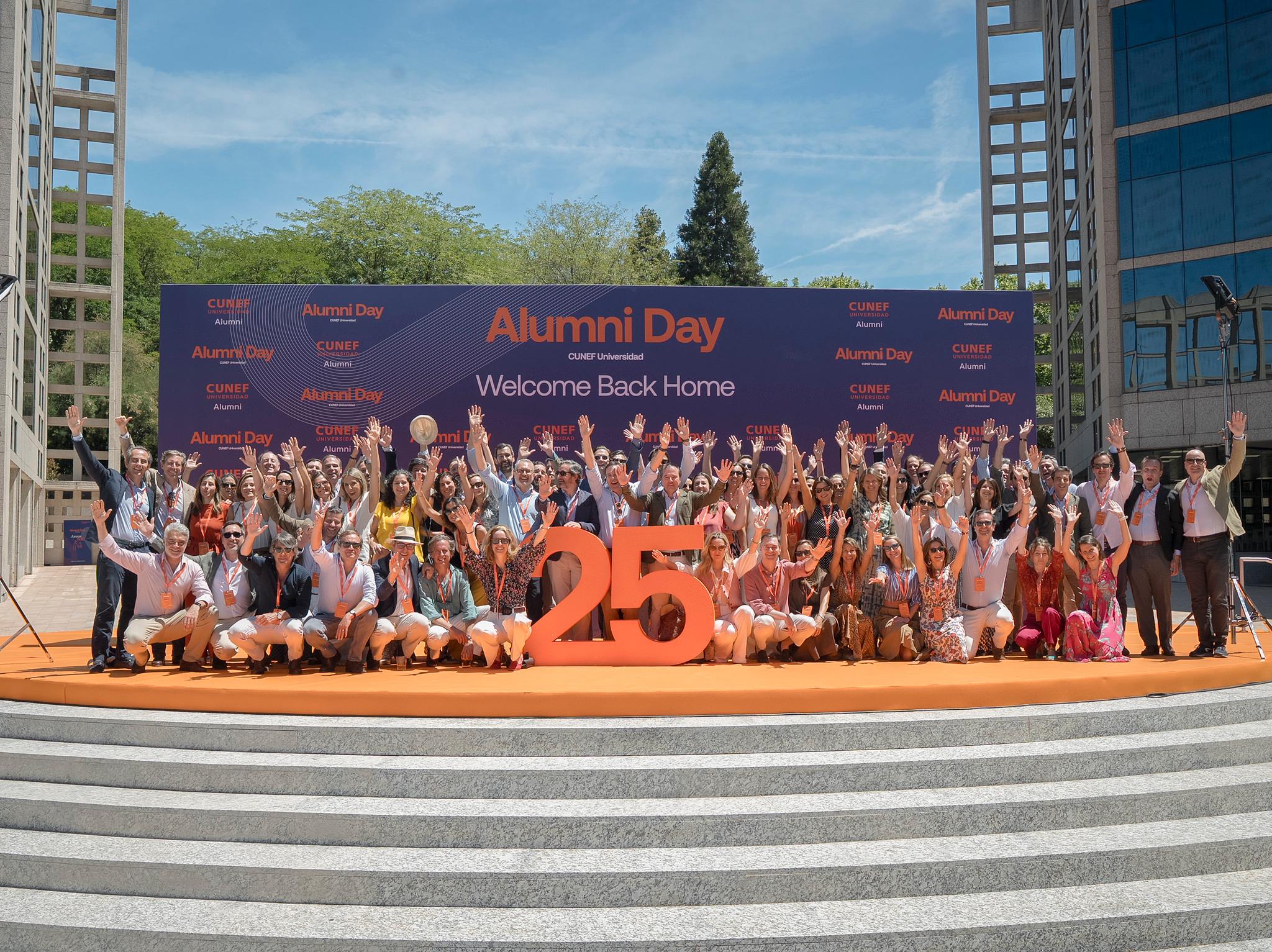 Celebración del Alumni Day con un grupo de personas levantando las manos frente a un gran cartel con el número 25.