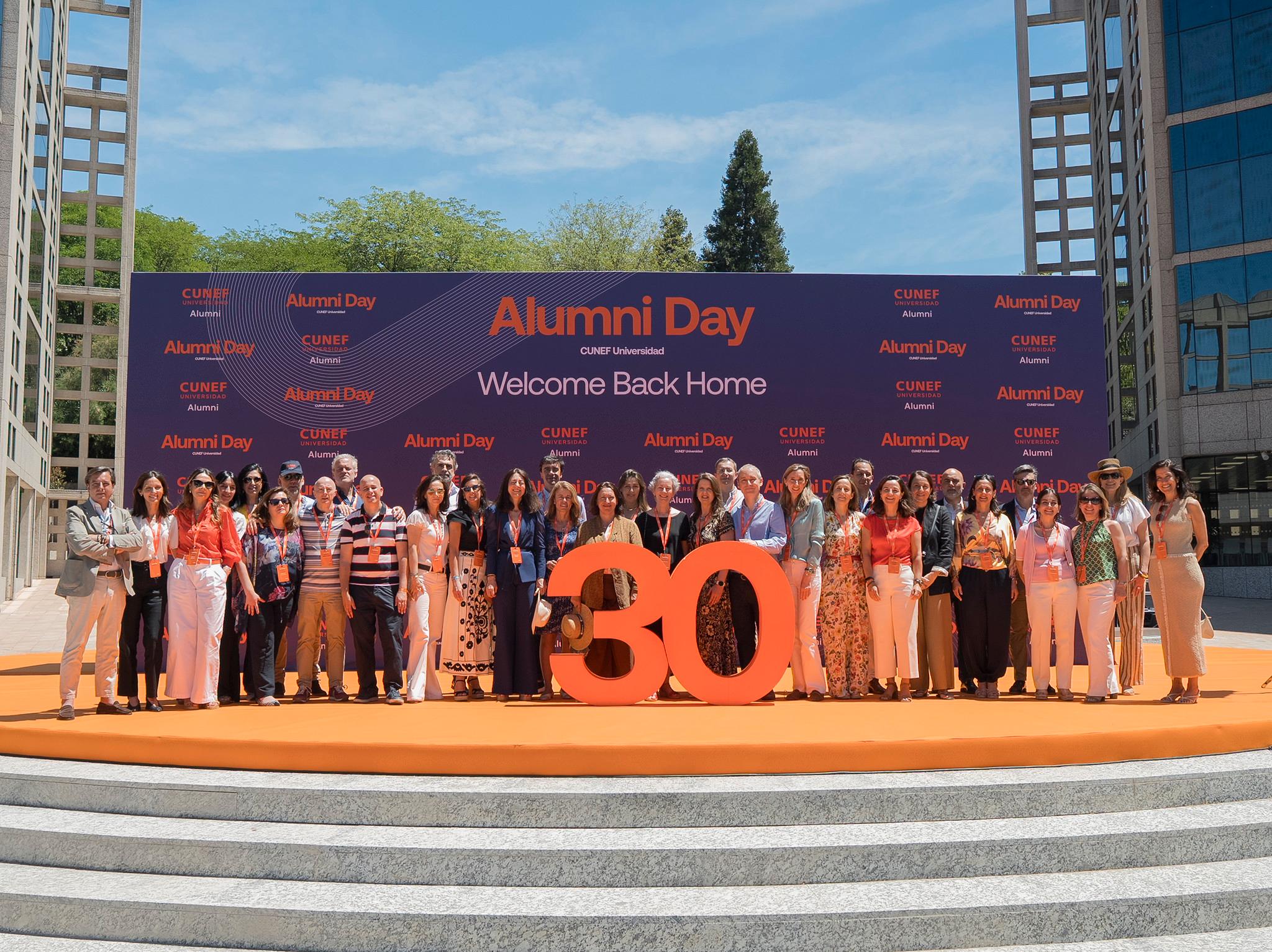 Un grupo grande de personas posando en un evento con un fondo que dice 'Alumni Day' y un número 30 destacado.