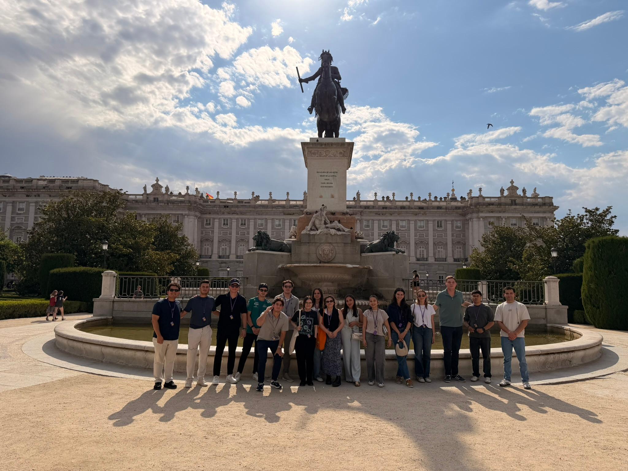Grupo de personas posando frente a una estatua en un parque con un fondo arquitectónico.