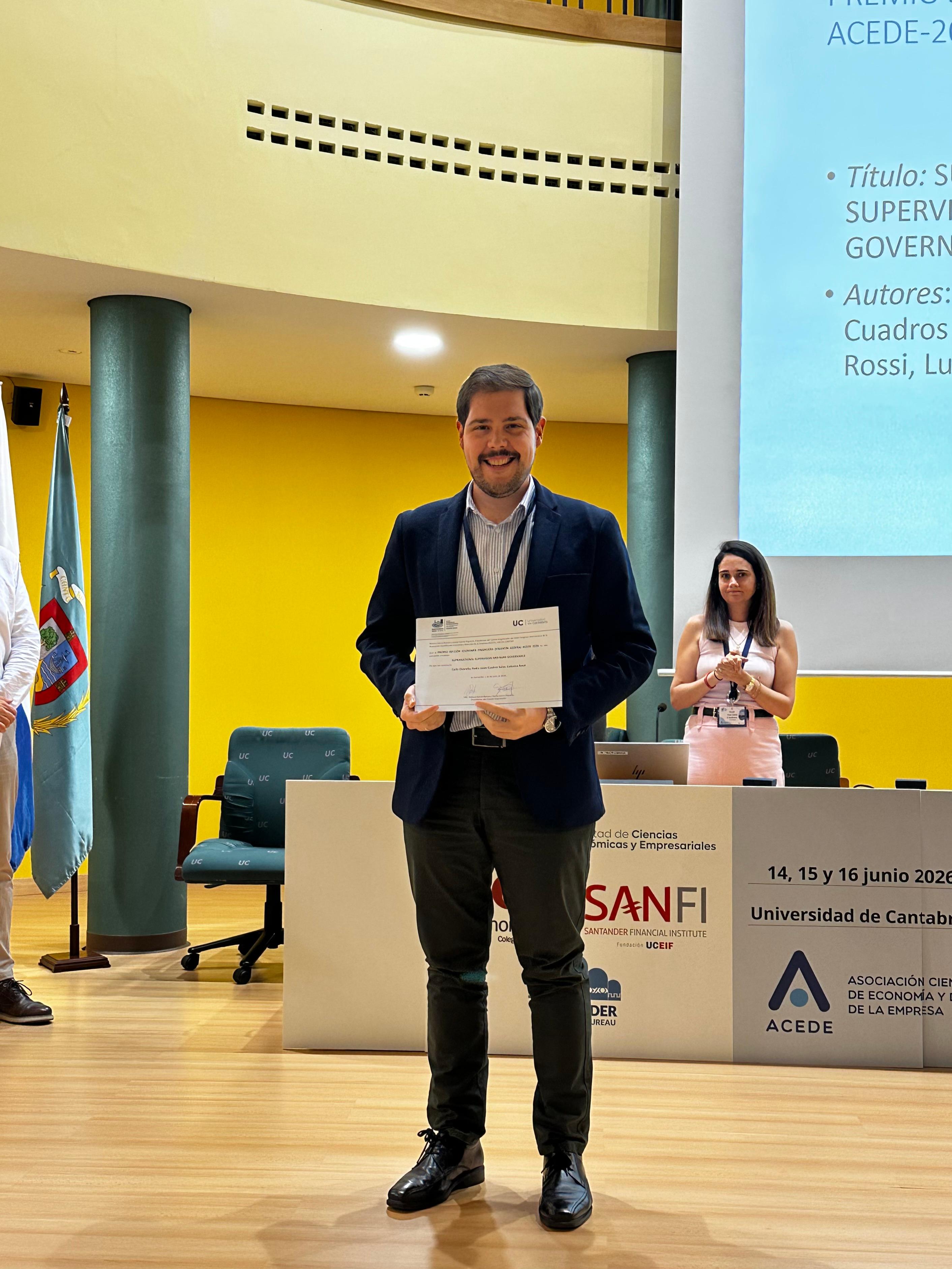 Un hombre sonriente sostiene un certificado en un congreso académico, mientras una mujer lo observa al fondo.
