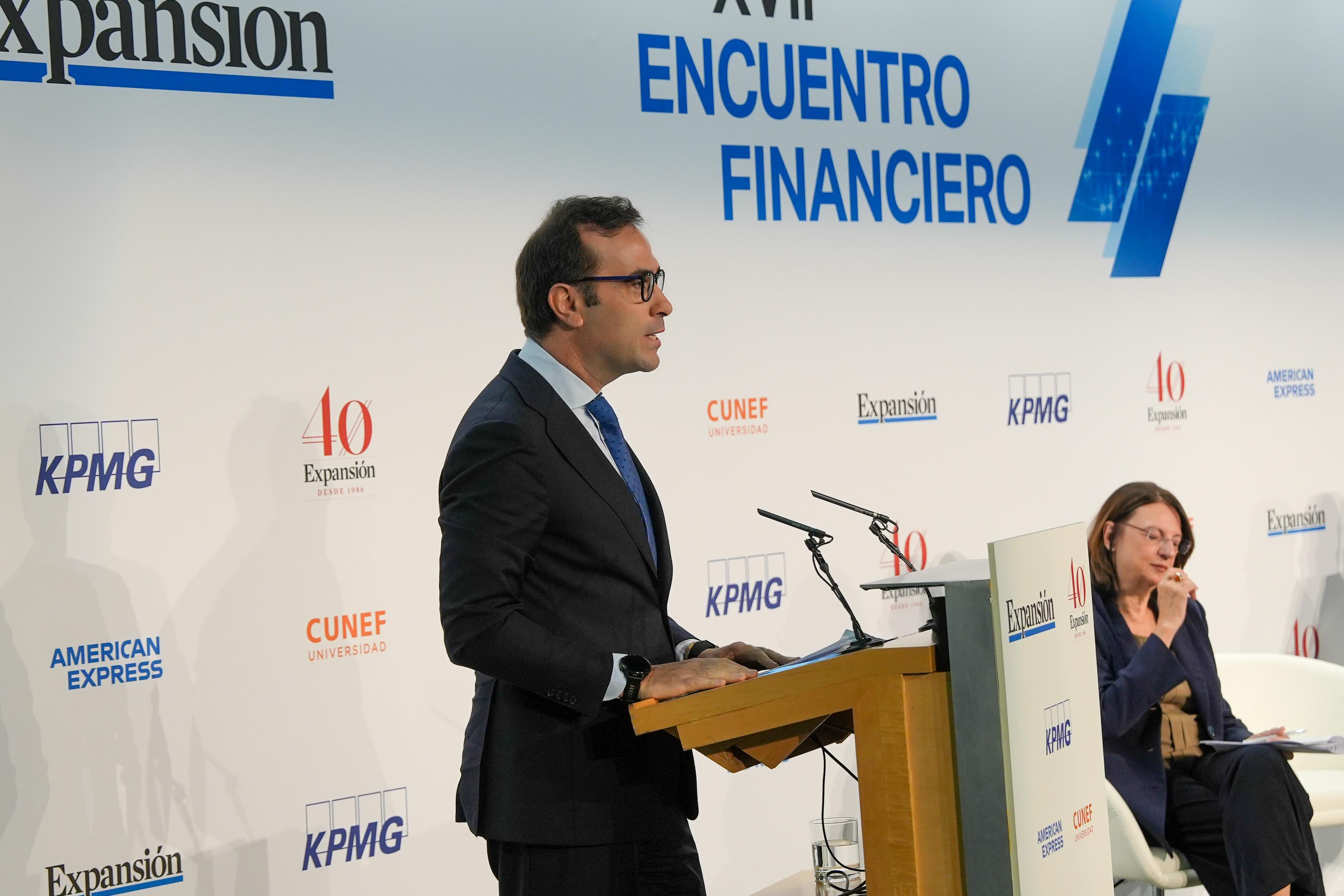 Un hombre está hablando en un podio durante un evento financiero en presencia de una mujer sentada al lado.